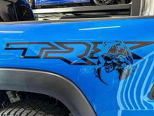 Load image into Gallery viewer, RAM TRX bedside decal on bright blue truck panel, featuring OEM logo and custom T-Rex dinosaur graphic. Black vinyl design contrasts against vibrant paint, showcasing unique Raptor Eater logo add-on for RAM enthusiasts.