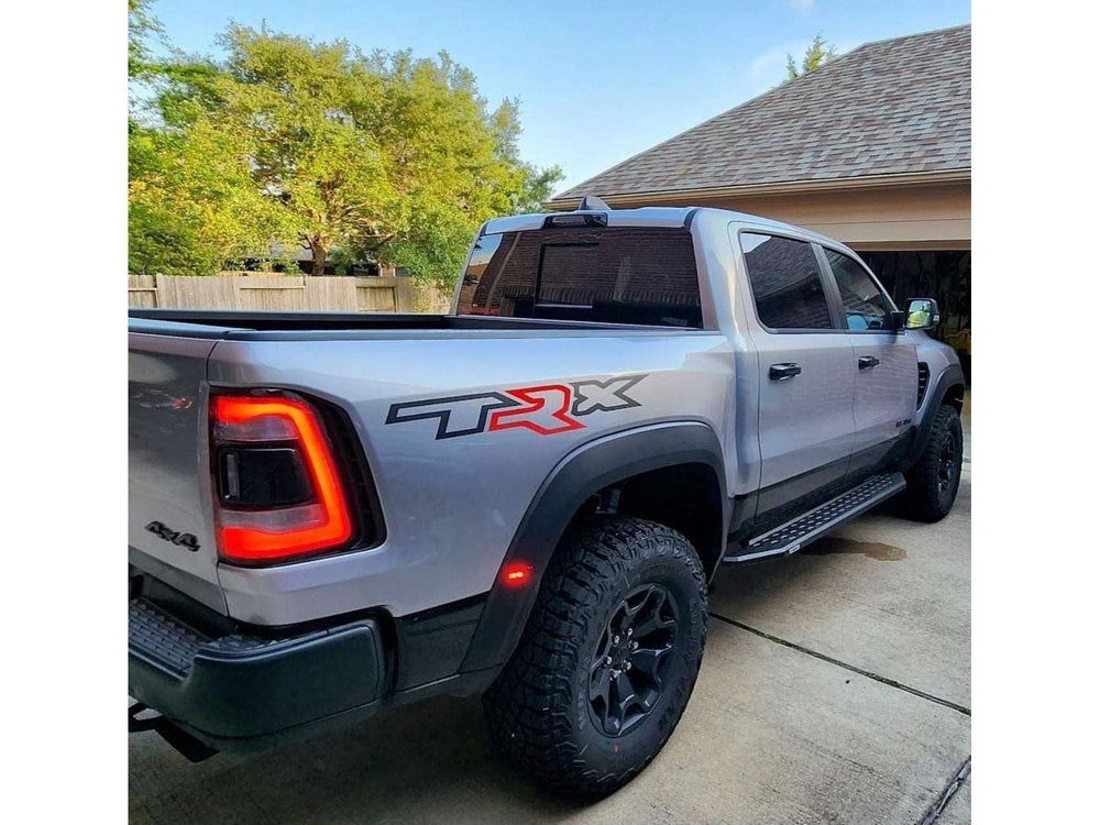 Silver RAM TRX pickup truck with customized bedside decal featuring TRX logo and colored R, parked in driveway next to house with trees in background. Showcases rugged off-road tires and distinctive rear tail lights.