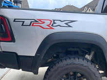 Load image into Gallery viewer, Ram TRX bedside decal on white truck, featuring black TRX logo with red R, showcasing custom vinyl design on vehicle&#39;s side panel above off-road tire