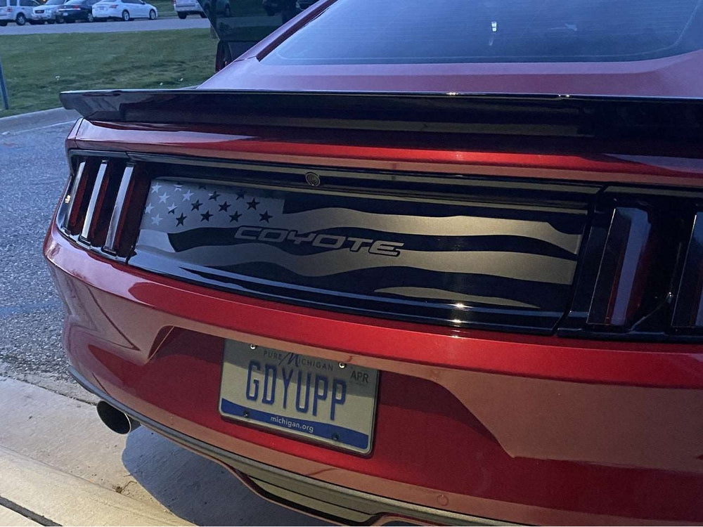 Red S550 Mustang rear view showcasing custom decklid decal featuring American flag design with Coyote text, enhancing the car's sleek appearance and patriotic styling. License plate visible, parked in outdoor setting.