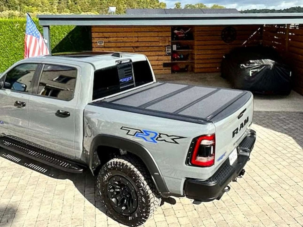 RAM TRX pickup truck with custom bedside decals parked in driveway, featuring tri-fold tonneau cover and off-road tires. American flag visible in background near wooden garage structure.