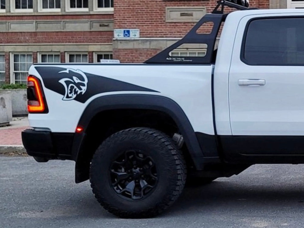 White RAM TRX pickup truck with black Hellcat logo decal on bedside, parked on street. Visible are large off-road tires, black fender flares, and distinctive RAM taillights. Building facade in background.