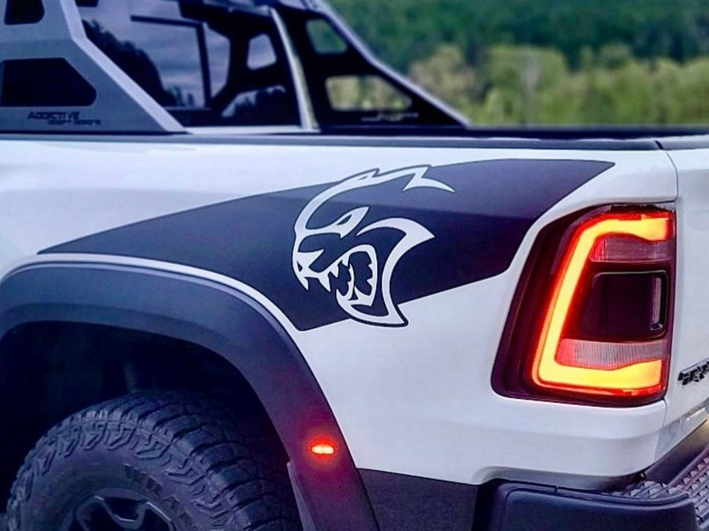RAM TRX truck side view showcasing custom Hellcat logo bedside decal, taillight, and off-road tire. White exterior with black graphic and illuminated red taillight visible against blurred natural background.