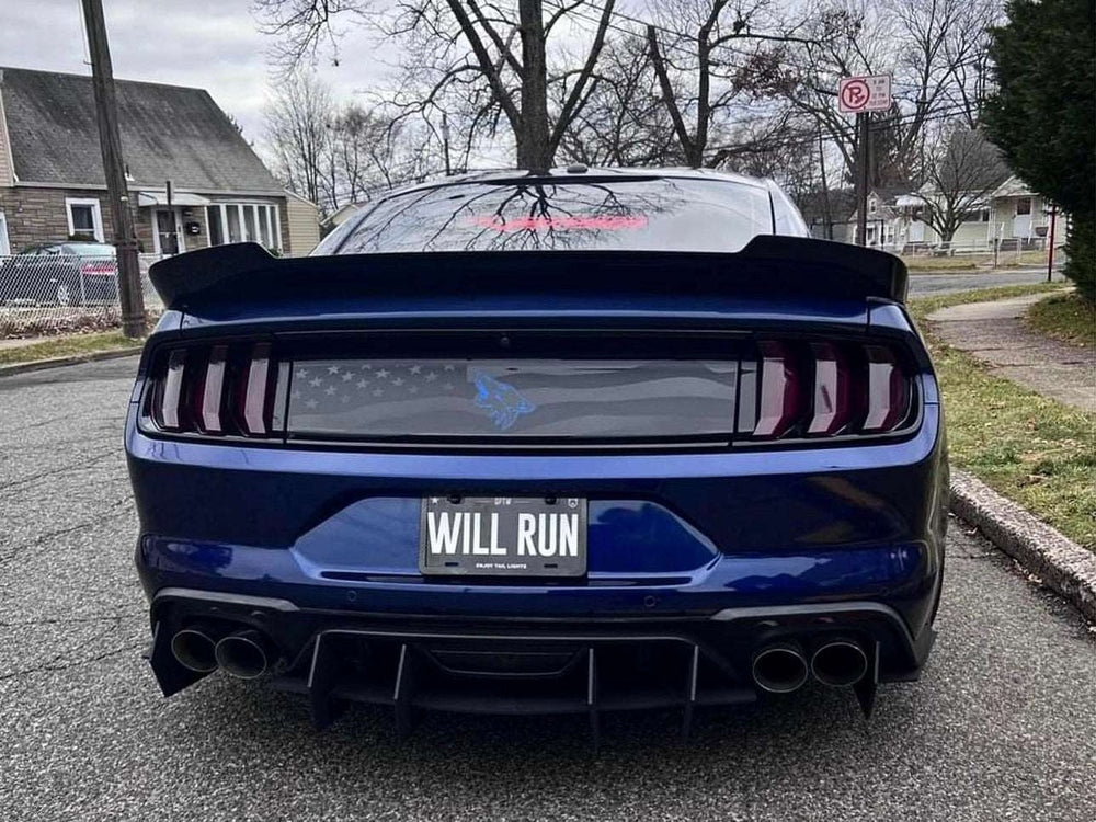 Rear view of blue Ford Mustang S550 with American flag decklid decal and Coyote logo, showcasing custom license plate "WILL RUN" and quad exhaust tips on suburban street