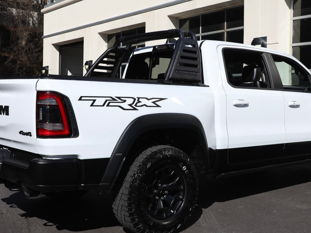 White RAM TRX pickup truck with black bedside decal, showcasing OEM-style design. Truck features off-road tires, black trim, and a sport bar in the bed, parked in front of a building with garage doors.