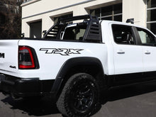 Load image into Gallery viewer, White RAM TRX pickup truck with black bedside decal, showcasing OEM-style design. Truck features off-road tires, black trim, and a sport bar in the bed, parked in front of a building with garage doors.