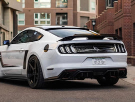 White Ford Mustang S550 parked on street, rear view showcasing sleek design, black wheels, and distinctive taillights. Urban brick buildings in background.