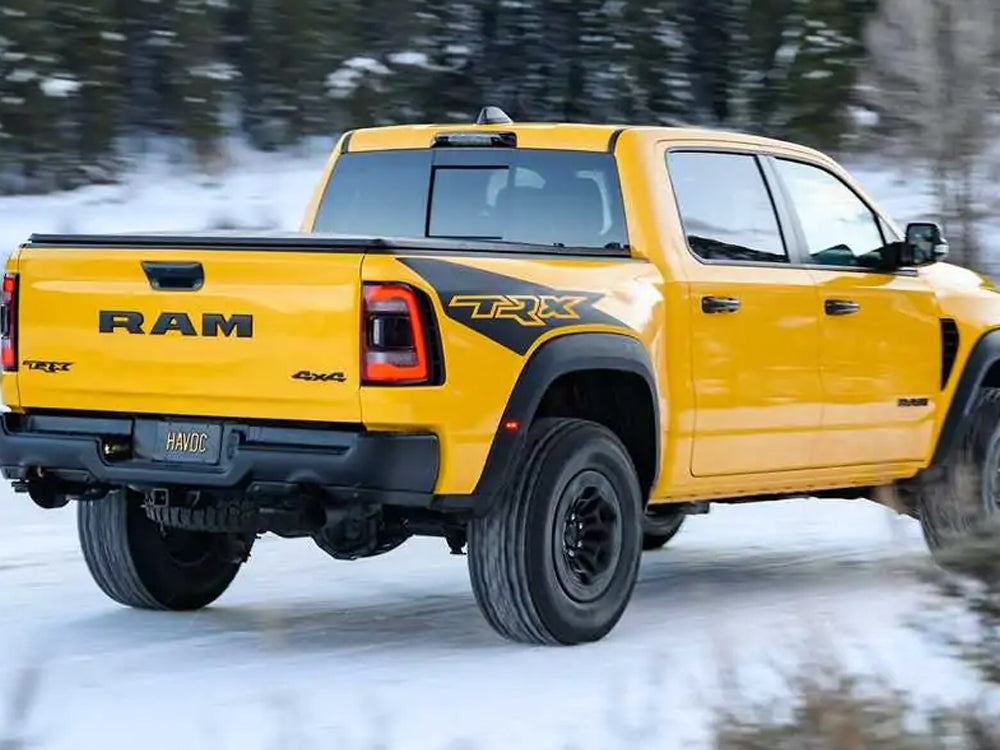 Yellow RAM TRX pickup truck driving on snowy road, showcasing bedside decal with TRX logo. Rugged off-road tires and distinctive RAM tailgate visible. Winter forest background highlights the truck's adventurous capabilities.