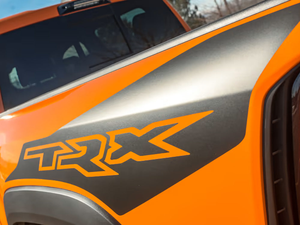 Close-up of RAM TRX truck bedside featuring orange Ignition Style decal with TRX logo cut out, revealing silver paint underneath. Orange and silver color scheme showcases the striking contrast and custom appearance of the product.