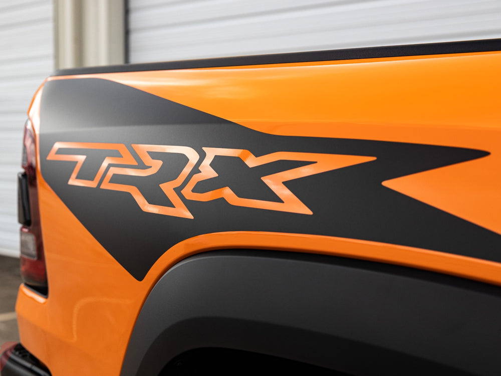 Close-up of RAM TRX truck bedside decal in black and orange, showcasing ignition style design on vibrant orange truck body against white background, highlighting custom vinyl graphics and OEM-style appearance