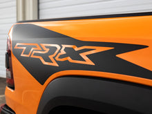 Load image into Gallery viewer, Close-up of RAM TRX truck bedside decal in black and orange, showcasing ignition style design on vibrant orange truck body against white background, highlighting custom vinyl graphics and OEM-style appearance