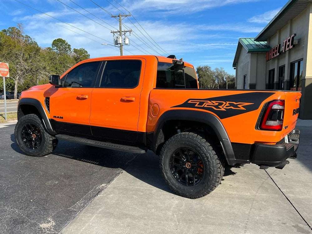 Bright orange RAM TRX pickup truck parked outside, showcasing custom black bedside decals with TRX logo. Large off-road tires and lifted suspension visible. Blue sky and building in background.