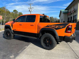 Bright orange RAM TRX pickup truck parked outside, showcasing custom black bedside decals with TRX logo. Large off-road tires and lifted suspension visible. Blue sky and building in background.