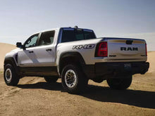 Load image into Gallery viewer, RAM RHO pickup truck in silver and black, parked on desert terrain with blue sky background. Side view showing bedside decal and rugged off-road tires.