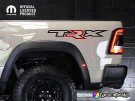 RAM TRX bedside decal on light-colored truck, featuring black 'T' and 'X' with red 'R'. Official licensed product logo visible. Rugged tire and tail light visible, showcasing truck's off-road capabilities.