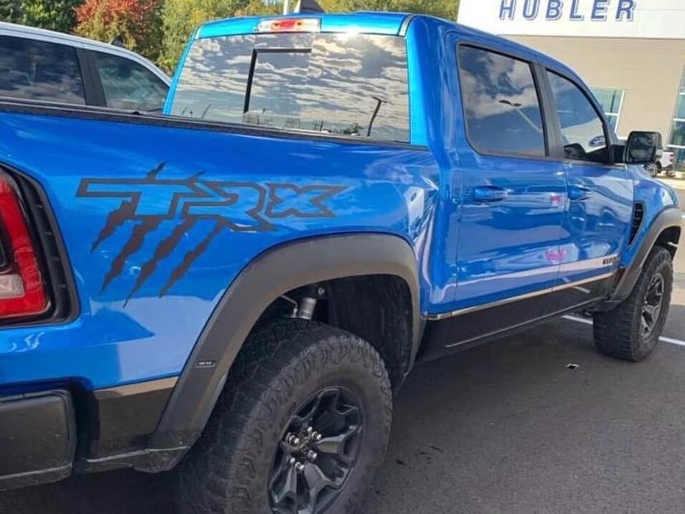 Blue RAM TRX pickup truck with custom bedside decal featuring TRX logo and claw marks, parked in front of a building with "HUBLER" sign visible.