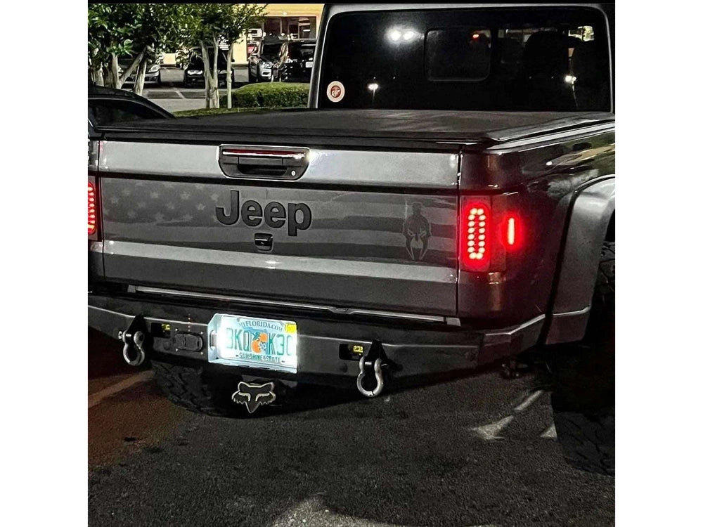 Jeep Gladiator tailgate with American flag and Gladiator logo decal at night, showcasing LED taillights and custom rear bumper with tow hooks, parked in a lot with trees visible in background
