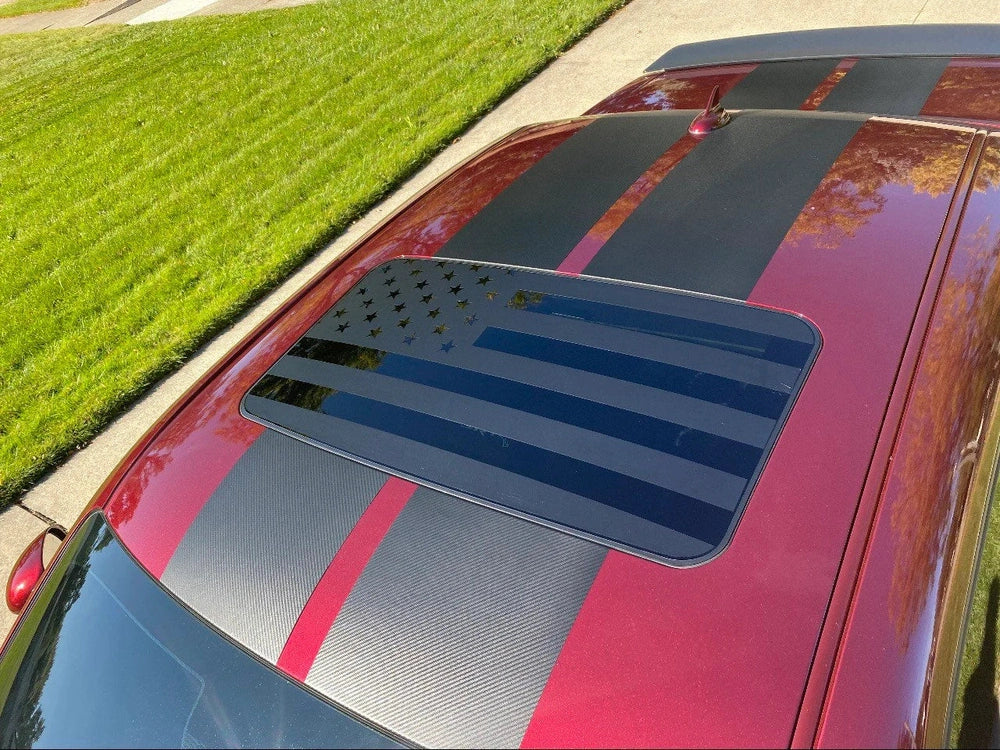 Red Dodge Challenger with racing stripes and American flag sunroof decal, parked on grass next to pavement, showcasing custom vinyl application for vehicle customization