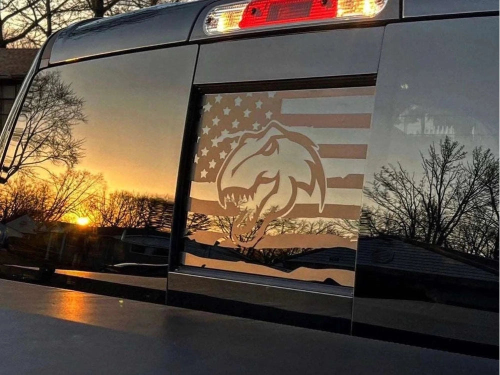 Ram truck rear sliding window with TRX Dino logo decal and American flag design, illuminated by sunset glow, reflecting trees and sky