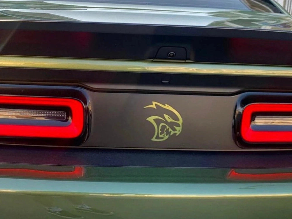 Dodge Challenger rear view with taillight divider decal featuring gold Hellcat logo between red LED taillights on dark panel, showcasing custom automotive styling and branding