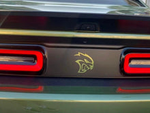 Load image into Gallery viewer, Dodge Challenger rear view with taillight divider decal featuring gold Hellcat logo between red LED taillights on dark panel, showcasing custom automotive styling and branding