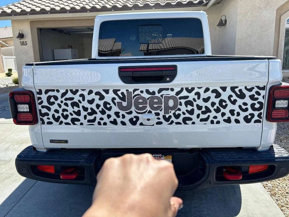 Jeep Gladiator tailgate with cheetah print decal, showcasing black spotted pattern on white background. Jeep logo visible in center. Rear view of truck parked in driveway.