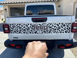 Jeep Gladiator tailgate with cheetah print decal, showcasing black spotted pattern on white background. Jeep logo visible in center. Rear view of truck parked in driveway.