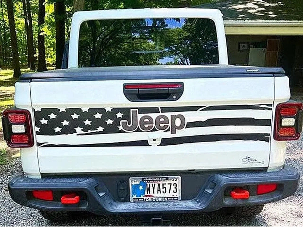 White Jeep Gladiator tailgate with American flag-inspired decal featuring stars, stripes, and Jeep logo, parked in a wooded area. Custom vinyl design showcases patriotic style on truck's rear.