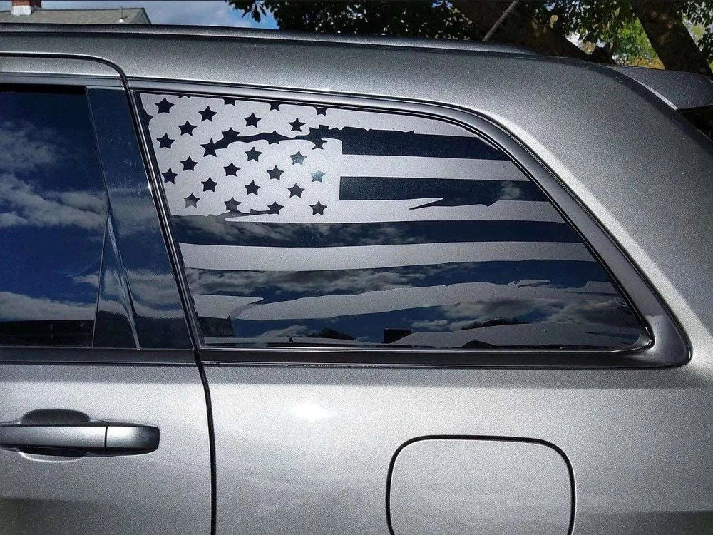 Jeep Grand Cherokee with American flag decal on quarter window, showcasing patriotic custom styling for 2011-2021 models. Silver vehicle exterior reflecting sky, demonstrating the sleek appearance of the matte black vinyl decal against the car's paint.