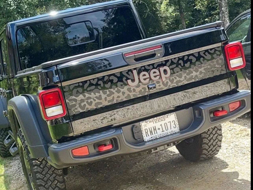 Jeep Gladiator tailgate with cheetah print decal, showcasing custom design on black vehicle exterior. Rugged off-road tires visible, parked in wooded area. Highlighting unique and stylish aftermarket modification for Jeep enthusiasts.